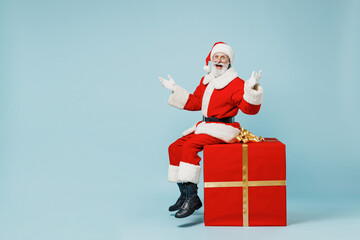 Full body old Santa Claus man in Christmas hat red suit clothes sitting on big gift present box spread arms isolated on plain blue background studio Happy New Year 2022 merry ho x-mas holiday concept