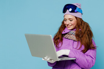 Skier smiling happy woman in warm purple padded windbreaker jacket ski goggles mask spend extreme weekend in mountains hold use work on laptop pc computer isolated on plain blue background studio