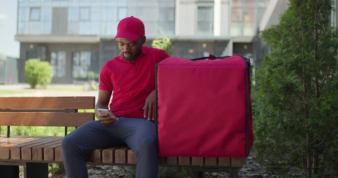 Black african delivery man sit on bench with thermo bag outdoors. courier carrying food and drinks order on city street. Male in red uniform waiting for clients, using smartphone, having rest