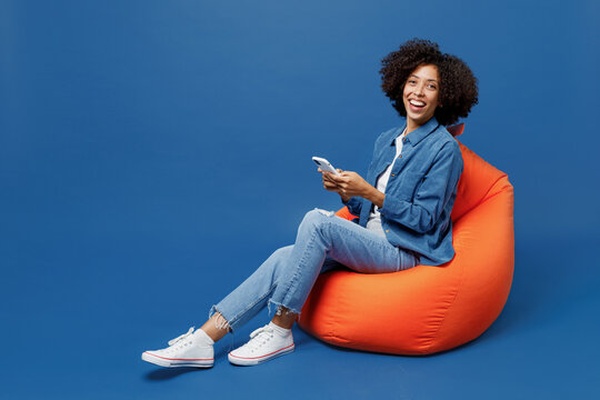 Full Body Young Smiling Cheerful Student Fun Happy Black Woman In Casual Clothes Shirt White T-shirt Sit In Bag Chair Hold Use Mobile Cell Phone Isolated On Plain Dark Blue Background Studio Portrait