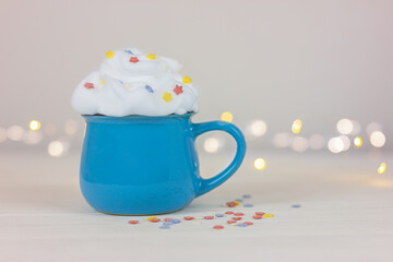 blue cup with a drink, decorated with whipped cream, on a white table against a background of festive garland lights