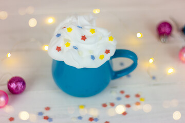 blue cup with a drink decorated with whipped cream and decorative stars on a white table next to christmas balls