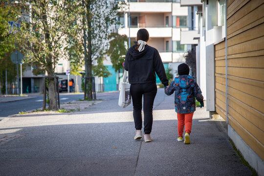 Back View Of A Young Woman And A Kid Walking On The Sidewalk On A Sunny Day