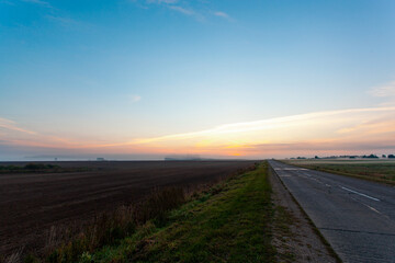 An empty asphalt road through the fields and forest in a thick fog at sunrise