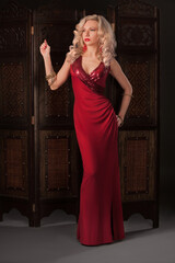 woman in red dress - sensual beautiful blonde model posing stands in red dress in a dark studio. Attractive sexy girl with long curly hair near retro background folding screen, fur. Blurred details