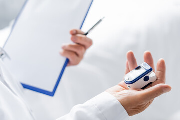 Cropped view of pulse oximeter and blurred clipboard in hand of doctor in clinic