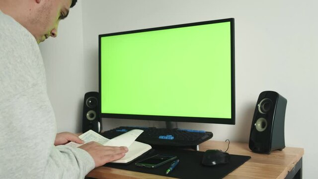 Over His Shoulder Is A Picture Of A Guy Working From Home On A Computer On A Desk, Looking At A Green Display Screen And Making Notes In His Notebook. Green Screen. High quality 4k footage
