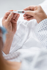 Cropped view of senior patient holding electronic thermometer on bed in clinic