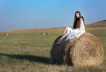 Naklejka na ściany i meble Beautiful long haired girl in big hat and simple boho styled dress in field with stacks of hay. Portrait of young woman with neutral makeup and curly hair 