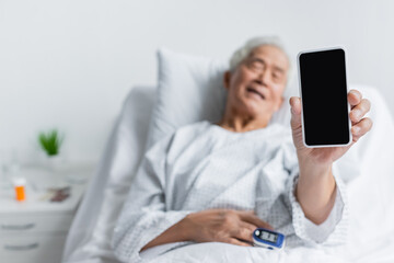 Smartphone in hand of blurred asian patient with pulse oximeter in hospital ward