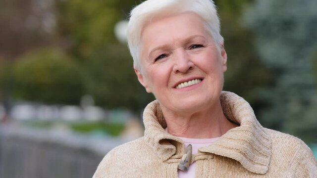 Close up portrait happy attractive benevolent woman of retirement age. Calm smiling lady in casual clothes standing posing outdoors looking at camera fixing gray short hair stylish hairstyle with hand