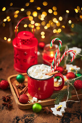 Red cup of hot cocoa with marshmallows and candy canes on wooden table with christmas tree and glowing garland for christmas