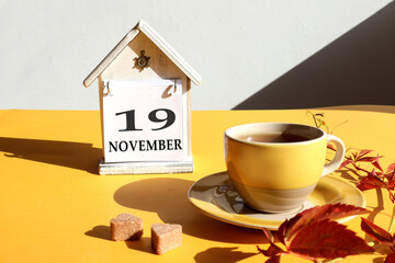 Calendar for November 19: a cup of tea with branches of maiden grapes against the background of a decorative house with the name of the month in English and the number 19, gray background