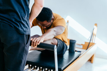 Rear view of instructor corrects and controls the Pilates exercise that his african american male student is doing on Reformer bed in health center or studio pilates.