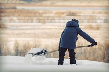 A teenager with a snow shovel cleans a path among snowdrifts on the background of nature. Snow...