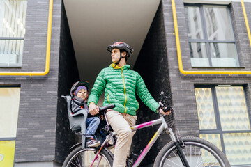 Little boy in the seat bicycle behind father. Caucasian white man in sportswear and helmet with his little son riding with bike at city street, outdoor. Safe and child protection concept.