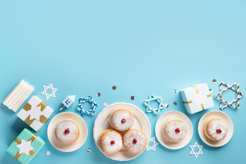 Jewish holiday Hanukkah concept - Hanukkah sweet doughnuts sufganiyot (traditional donuts) with powdered sugar and fruit jelly jam, gift boxes, spinnig driedel and candles on blue paper background.