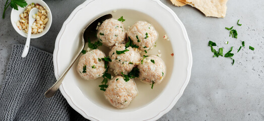 Homemade chicken matzo ball soup with parsley and garlic in simple white ceramic plate on a gray stone or concrete background. Traditional Jewish passover dish. Selective focus. Top view