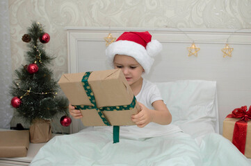 Obraz na płótnie Canvas Christmas morning, little girl in pajamas with gifts in bed against the background of Christmas tree. A happy, smiling child opens New Year's gift at home. The concept of holidays, New Year, Christmas