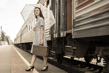 An elegant lady in a dress with an umbrella and a vintage suitcase gets off the train on the...