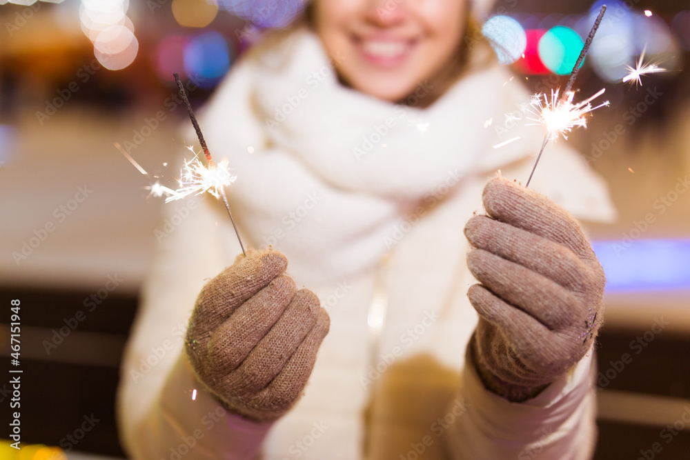 Wall mural Smiling young woman wearing winter knitted clothes holding sparkler outdoors over snow background. Christmas holidays.