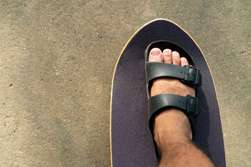 men's feet In sandals, stepping on a black Surf Skate on the sand in the park.