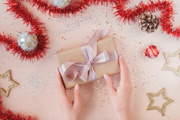 Female hands with natural manicure holding present in kraft paper with pink ribbon on soft beige background. Xmas composition. Flat lay. Happy holidays, New Year celebration and giving love concept. 