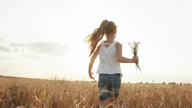 A girl in a white tunic runs to the sun across a wheat field with a bouquet of chamomile in her hand. Back view. Slow motion