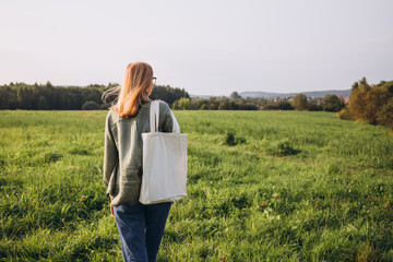 Young blonde woman with white cotton bag in her hands on nature background. Empty reusable canvas...