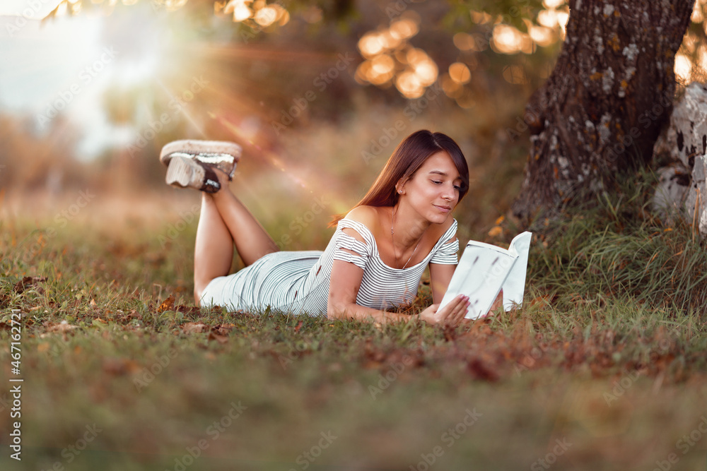 Poster shallow focus of a caucasian female with a white dress posing in the park and reading book