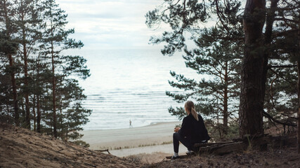 Fototapeta na wymiar Portrait of a Young Beautiful Blond Woman in a Romantic Nature Atmosphere. Girl is Dressed in Black and is Sitting Alone in a Forest. She is Looking at a Sea Landscape. Man Running in the Distance.