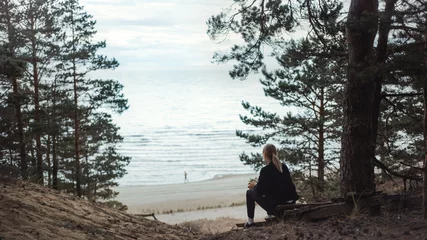 Foto auf Acrylglas Portrait of a Young Beautiful Blond Woman in a Romantic Nature Atmosphere. Girl is Dressed in Black and is Sitting Alone in a Forest. She is Looking at a Sea Landscape. Man Running in the Distance. © Gorodenkoff