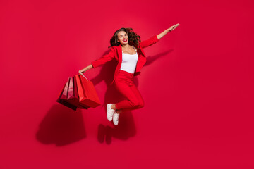 Full body photo of impressed brunette lady jump with bags wear suit shoes isolated on red color background