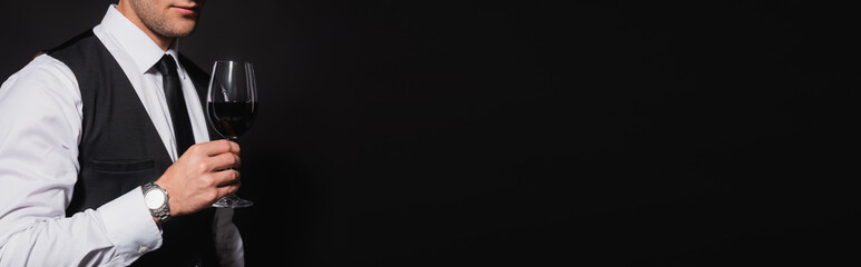 partial view of man in white shirt and vest holding glass of red wine isolated on black, banner