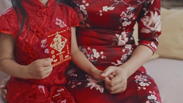 Mid-section close up shot of unrecognizable Chinese mother and little daughter in traditional dresses sitting on couch and holding hands. Girl is holding Lunar New Year postcard