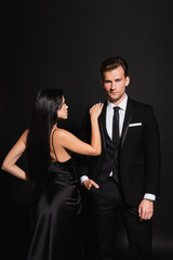 brunette woman in silk dress touching shoulder of elegant man standing with hand in pocket on black