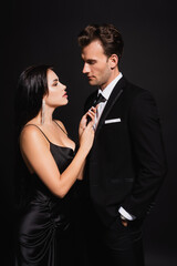 brunette woman seducing man in elegant suit and touching his tie isolated on black