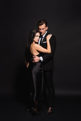full length view of man in suit hugging stylish woman in satin dress on black