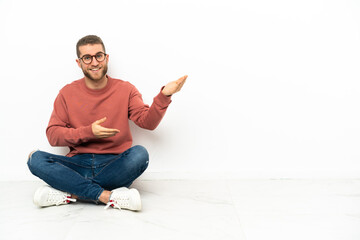 Young handsome man sitting on the floor extending hands to the side for inviting to come