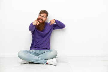 Fototapeta premium Young caucasian reddish man sitting on the floor isolated on white background points finger at you while smiling