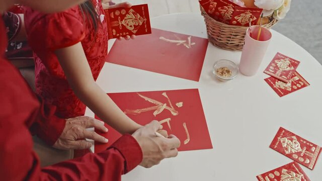 Handheld high angle shot of unrecognizable Asian father helping his little daughter drawing hanzi character on red sheet of paper while making postcard for Chinese New Year
