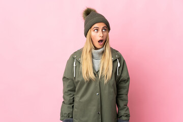 Young woman with winter hat isolated on pink background looking up and with surprised expression