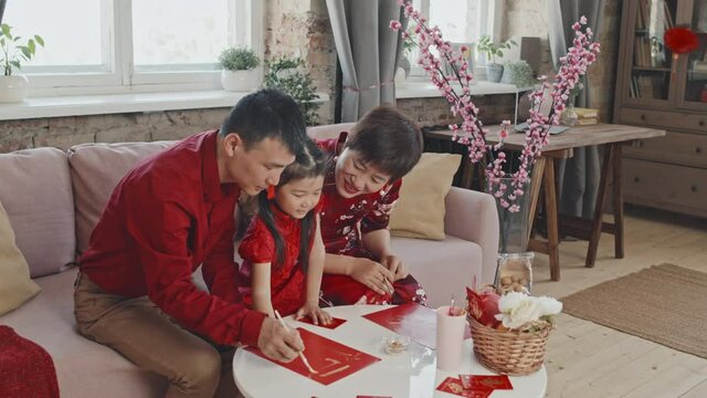 Medium shot of Asian parents helping their cute 5-year-old daughter in traditional dress making Chinese New Year postcard