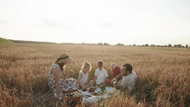 A friendly family with children sits around an impromptu table during a picnic in a wheat field on a sunny summer day. Slow motion