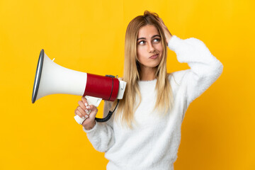 Young blonde woman isolated on yellow background holding a megaphone and having doubts