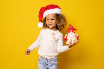 Beautiful smiling little girl in santa hat holding small santa over yellow background.