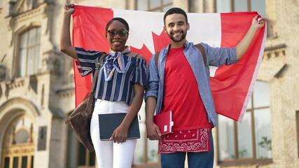 Portrait of happy African American woman and Middle Eastern man standing at college campus wrapping in Canadian flag smiling looking at camera. Proud intelligent young students posing outdoors - Powered by Adobe