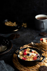Corn flakes with strawberry and blueberry in black bowls over dark background, top view.
