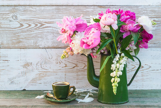 Beautiful greeting card or invitation for coffee break in retro style; Vintage cup of coffee and bouquet of pink peonies flowers in green coffee pot on rustic white paint wooden background; copy space