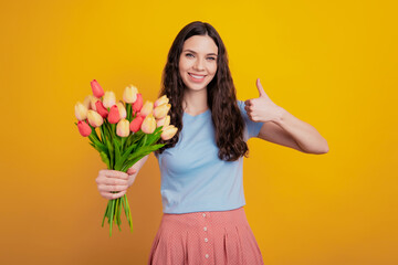 Obraz na płótnie Canvas Portrait of a happy young attractive girl holding tulips show thumb-up like isolated over yellow color background